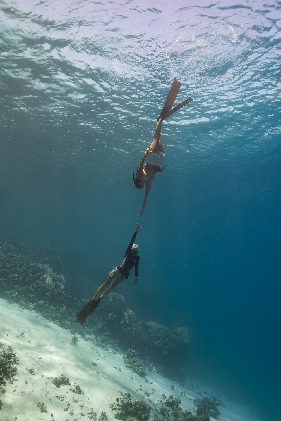 Two freedivers reaching toward each other above a sunlit coral reef